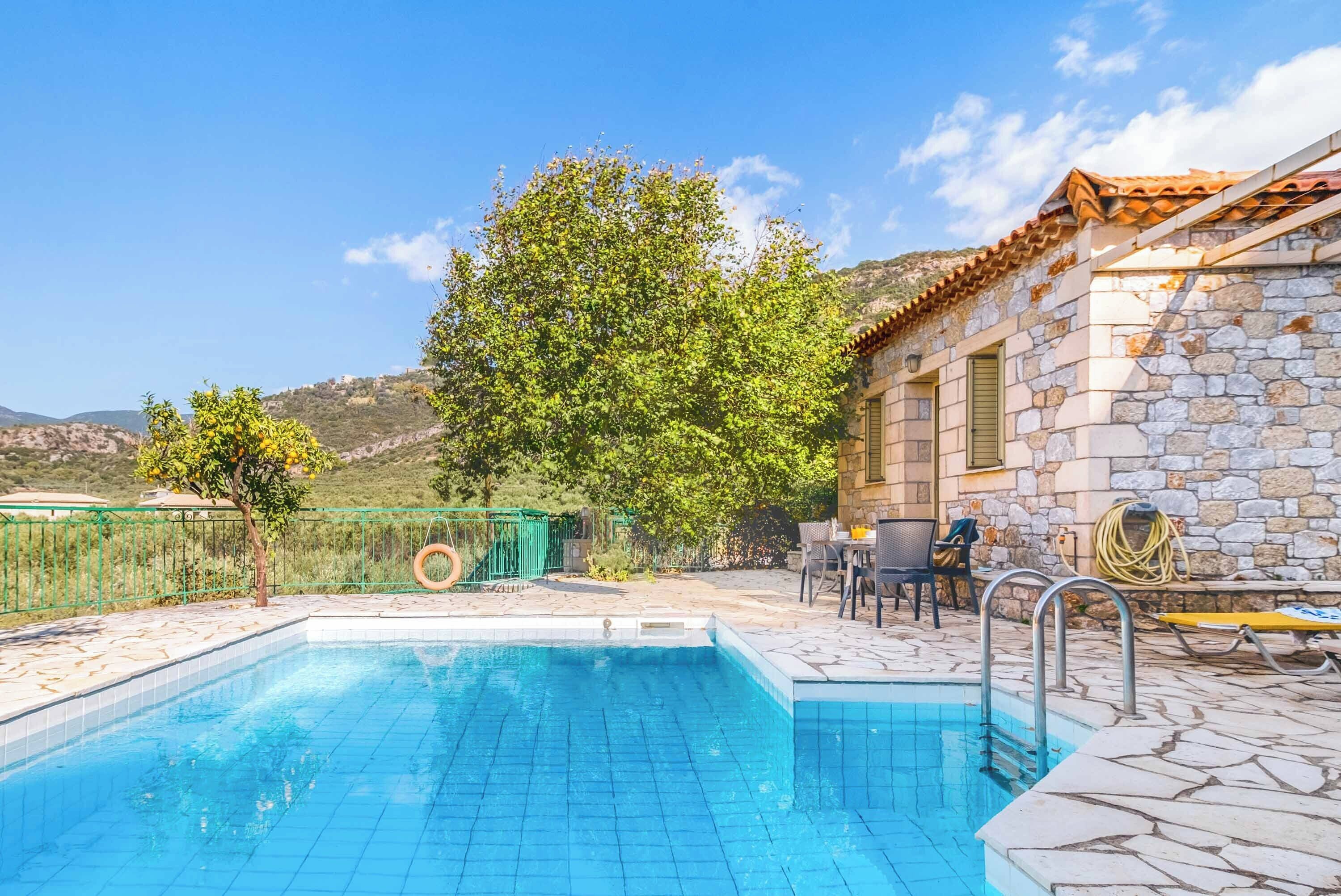 Una piscina con baldosas de piedra, un edificio de piedra con tejado a dos aguas, mobiliario de exterior y un árbol.