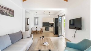 A modern living room with a sofa, a coffee table, and a TV. A kitchen is visible in the background with a dining area. - Villa Ira Lefkas - One Bedroom Villa, Sleeps 2 (Lefkada)
