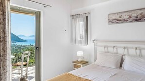 A bedroom with a bed, a nightstand, a lamp, a window with a view of the sea and mountains, and a painting on the wall. - Villa Ira Lefkas - One Bedroom Villa, Sleeps 2 (Lefkada)