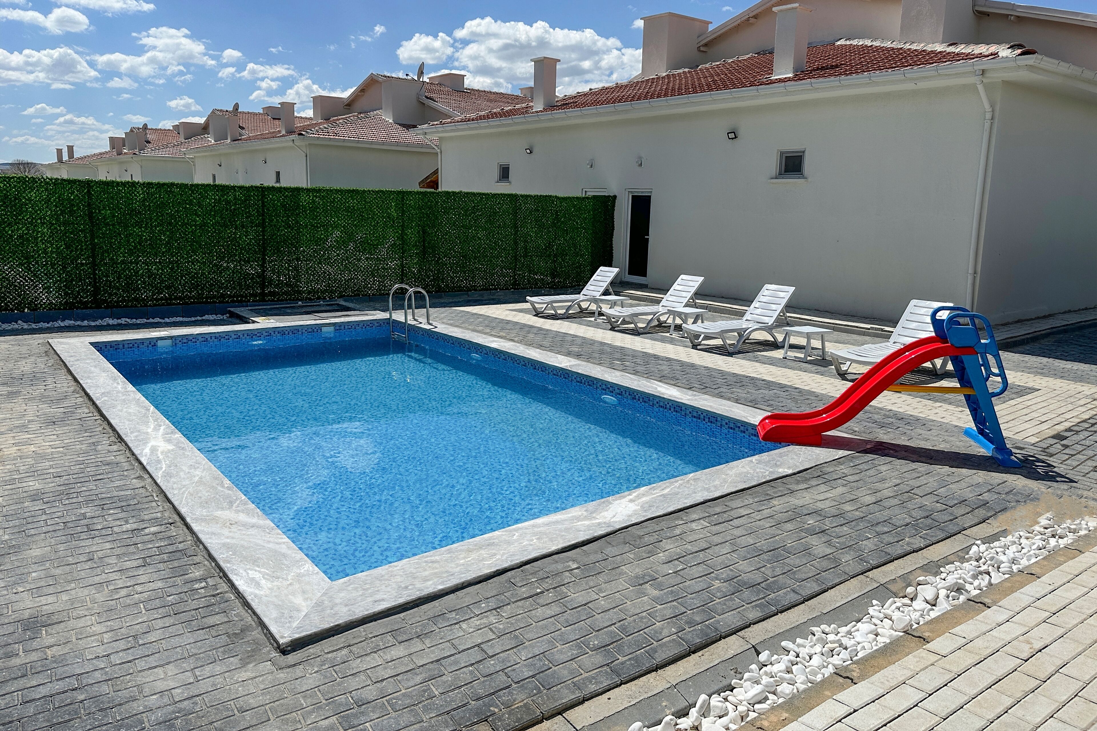 A rectangular swimming pool with a red slide, surrounded by lounge chairs and a green hedge.