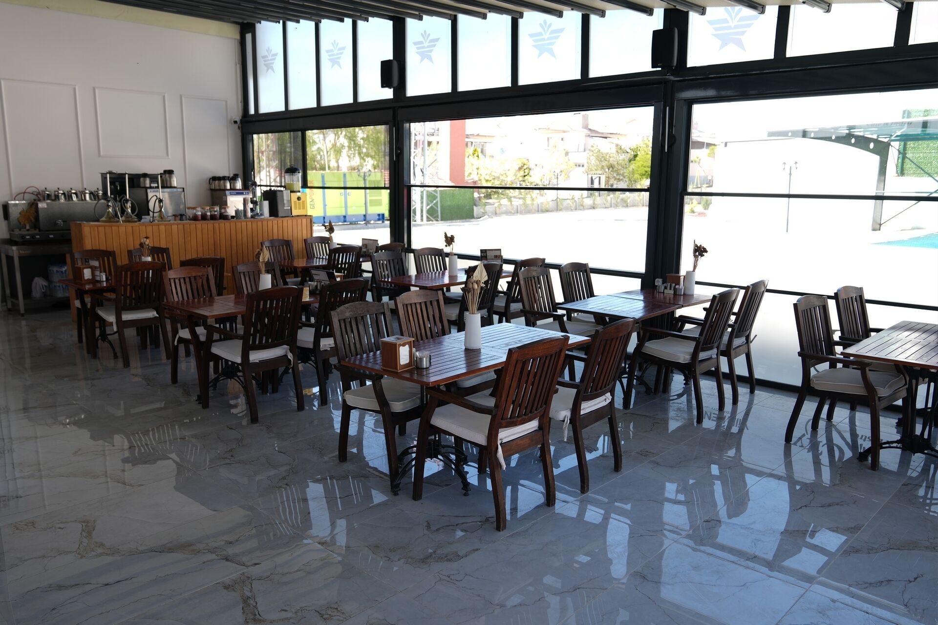 A spacious indoor dining area with wooden tables and chairs, large windows offering views of the outside, and a kitchen area in the background.