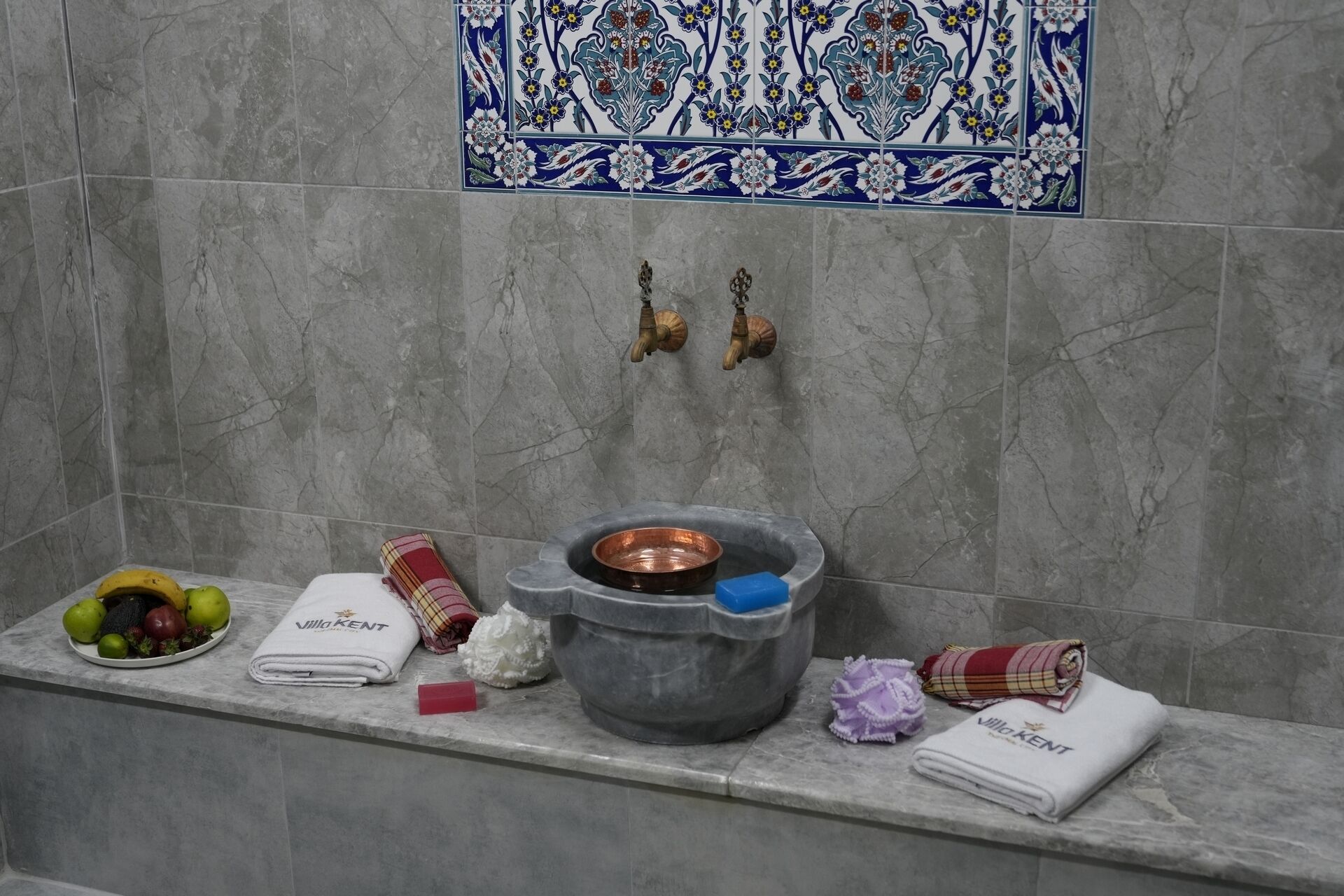 A bathroom with a marble wall, a decorative tile above the sink, and a shelf with towels, a basin, and toiletries.