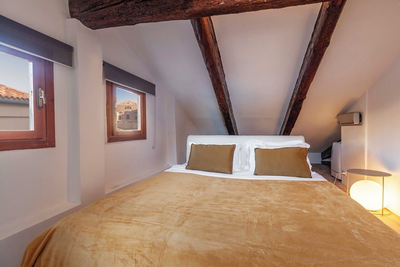 Une chambre avec un plafond en pente, un lit recouvert d’une couverture marron, deux oreillers, une lampe de chevet et une fenêtre donnant sur des immeubles.