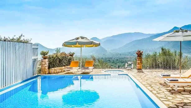 Ein Swimmingpool mit Liegestühlen und Sonnenschirmen, umgeben von Steinmauern und Blick auf Berge.