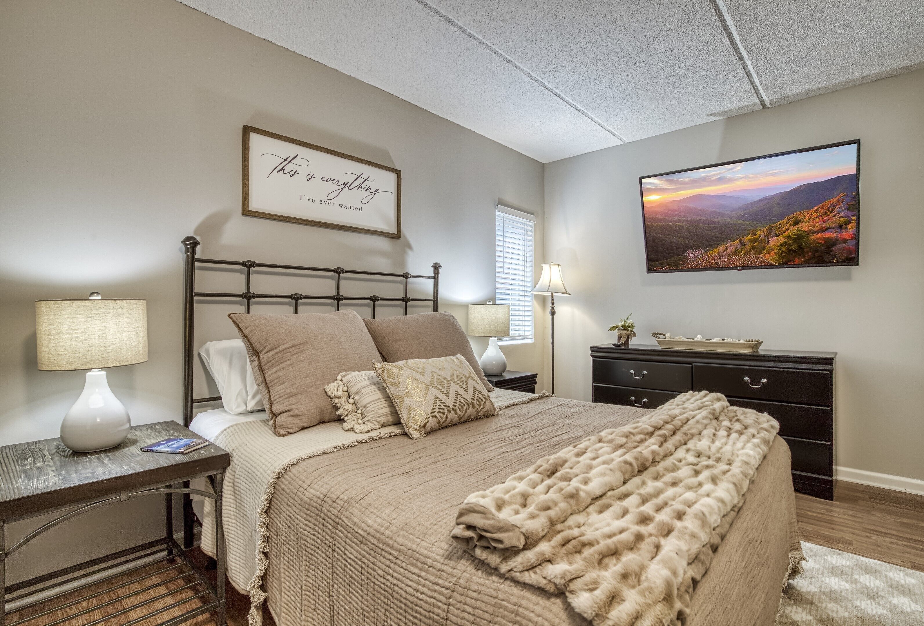 Une chambre à coucher avec un lit, deux tables de chevet avec des lampes, une commode, une citation encadrée au mur et un tableau représentant un paysage.