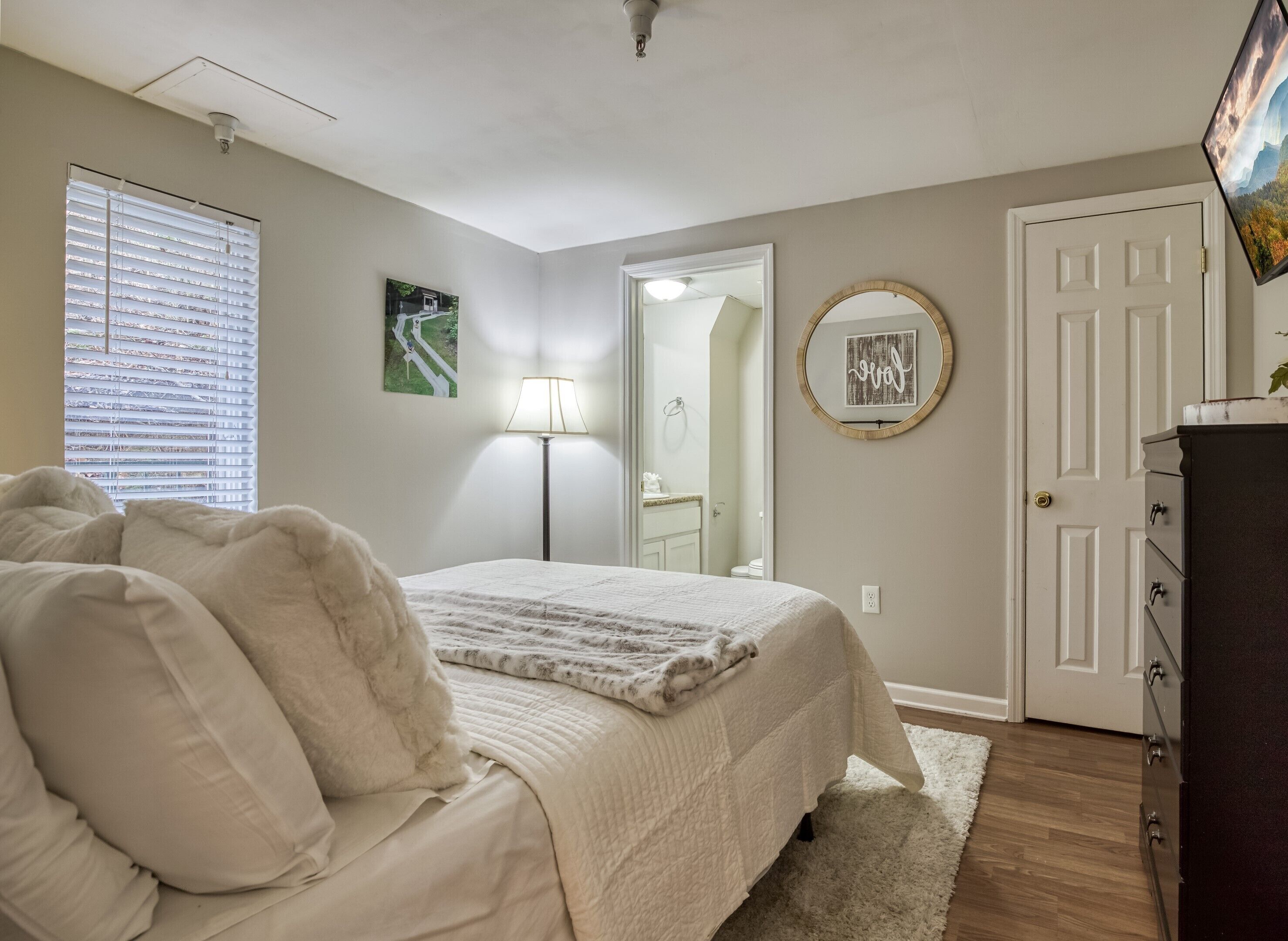 Een slaapkamer met een bed, een bedlamp, een dressoir, een deur en een raam met jaloezieën.