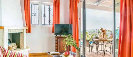 A living room with a sofa, a television, a table with a vase and fruit, and a view of the outdoors through a sliding glass door.
