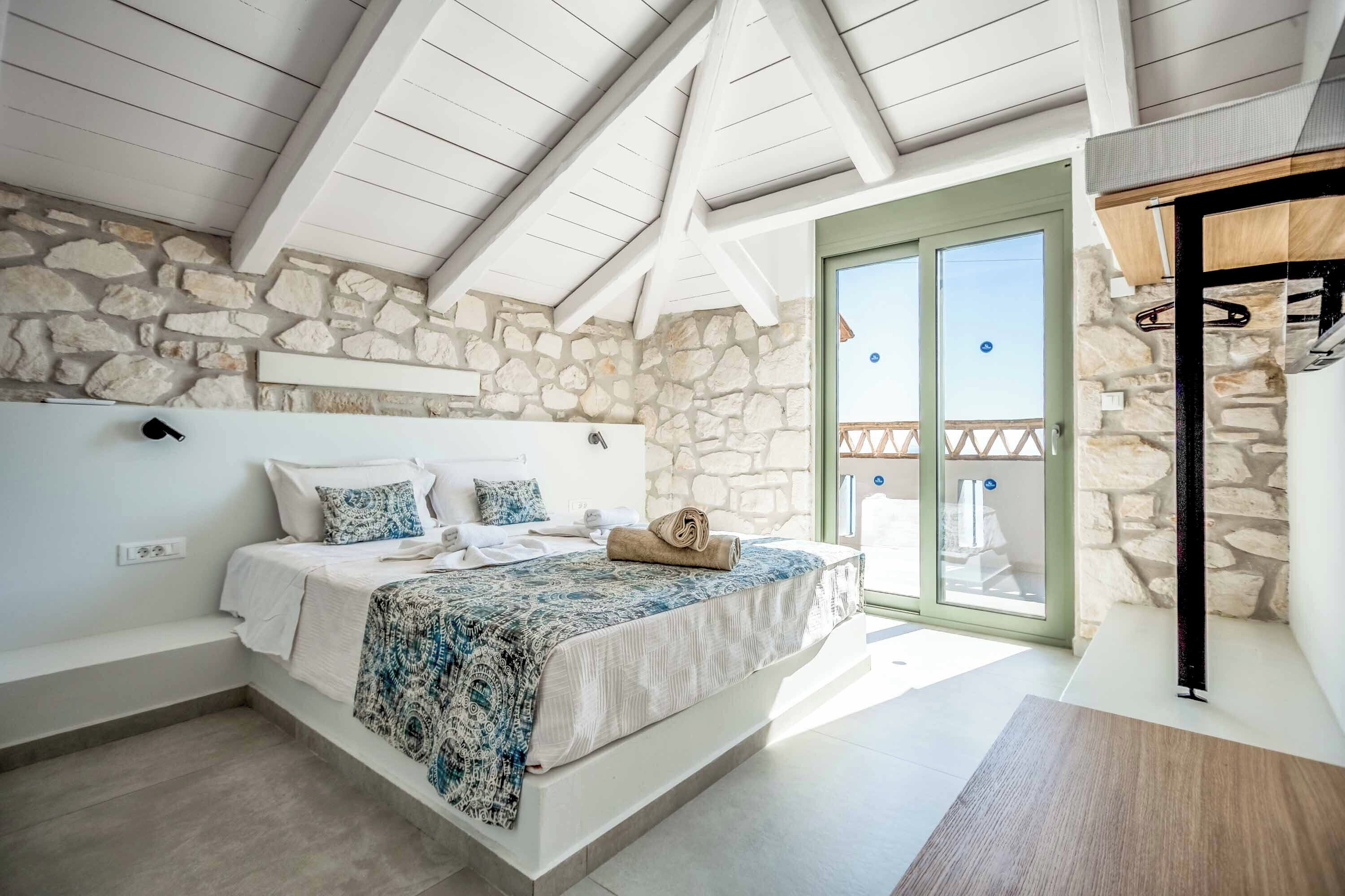 A bedroom with a stone wall, a bed with a patterned bedspread, a wooden bench, and a balcony with a view.
