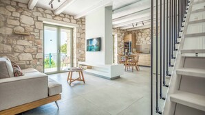 Un menjador modern amb una paret de pedra, un sofà, una zona de menjador i una cuina visible a través d'una porta oberta.