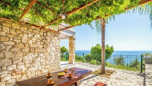 Mur de pedra amb taula i bancs de fusta, una pérgola amb vegetació i vista al mar.