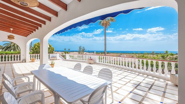 A covered outdoor patio with a white table and chairs, a view of the sea, and a palm tree.