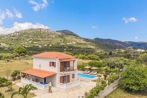 Ein zweistöckiges Haus mit Swimmingpool, umgeben von Grünflächen und Hügeln.