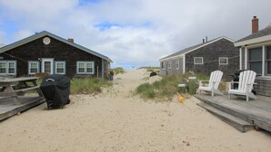 Ein Strandhaus mit einer Holzterrasse, einem überdachten Grill und einem Sandweg, der zum Haus führt.