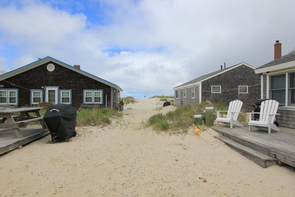 Een vakantiehuis aan het strand met houten huizen, een zandpad en strandstoelen.