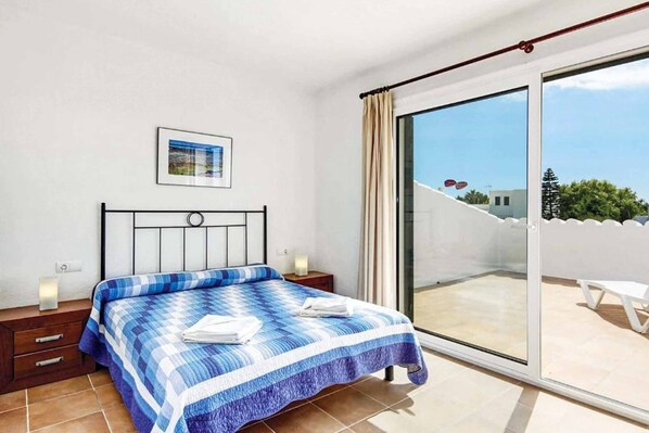 Un dormitorio con cama, mesita de noche, un cuadro enmarcado en la pared y un ventanal corredero que da a un balcón con una silla blanca.