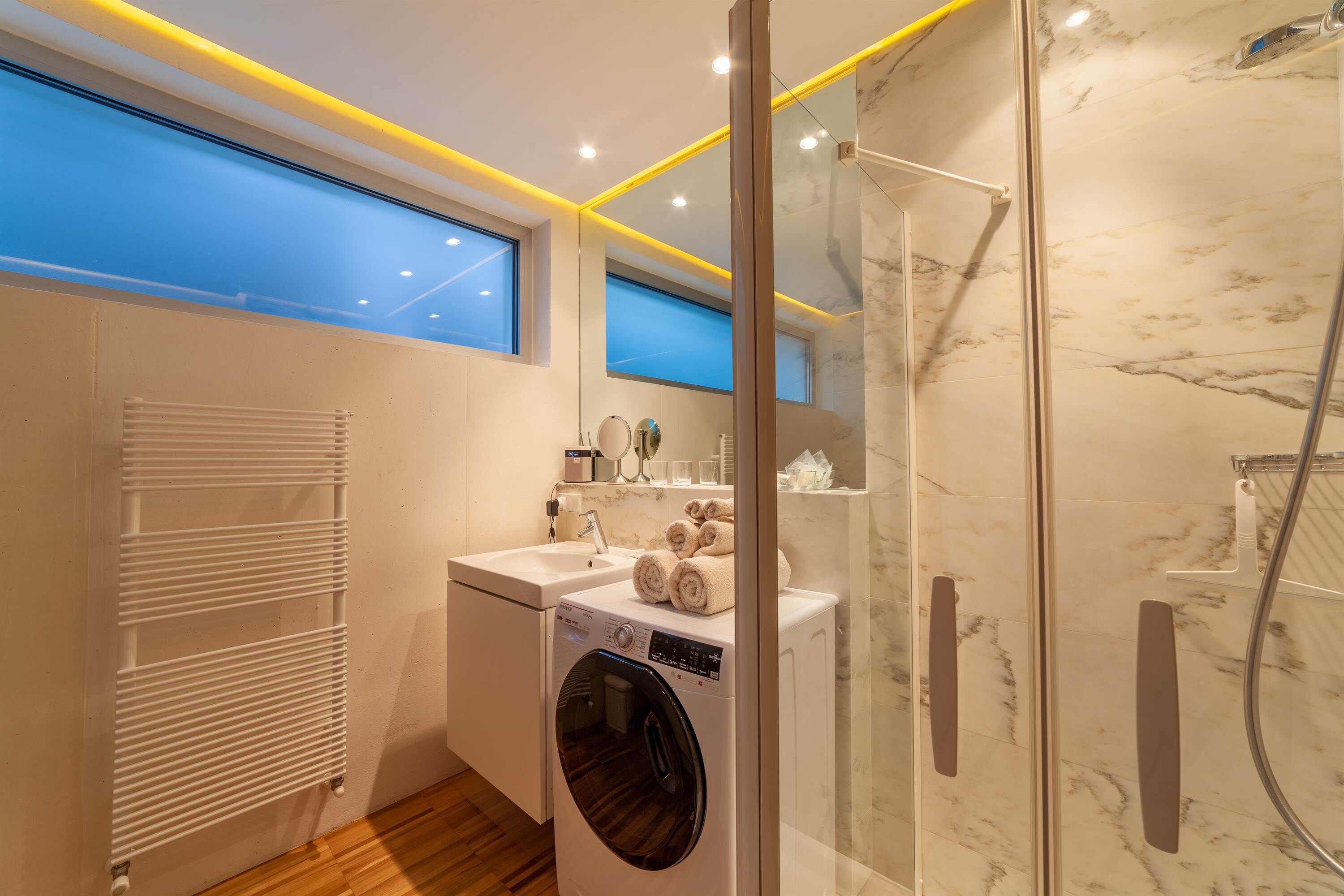 A modern bathroom with a glass-enclosed shower, a built-in washing machine, and a large mirror.