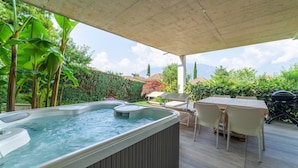 Un bain à remous sous une pergola en bois, entouré d’une terrasse avec une table et des chaises, et une végétation luxuriante en arrière-plan.