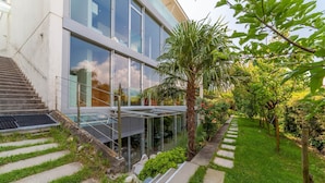 Un bâtiment moderne doté d’une façade en verre, d’un escalier menant à un espace extérieur couvert et d’un jardin bien entretenu avec un palmier et divers arbustes.