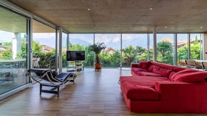 Un vaste espace de vie comprenant un canapé rouge, un fauteuil noir et un téléviseur posé sur un meuble. De grandes fenêtres offrent une vue sur la verdure et les bâtiments.