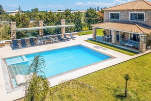 A swimming pool with lounge chairs, a covered patio, and a stone house with a tiled roof.