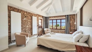 Une chambre avec un grand lit, deux fauteuils, un mur en pierre, une fenêtre avec des rideaux et une vue sur l’extérieur.