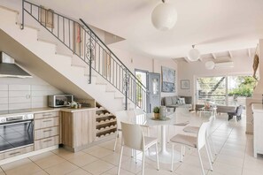 A modern kitchen with a built-in oven, a dining area with white chairs, a staircase with a metal railing, and a large window providing natural light.