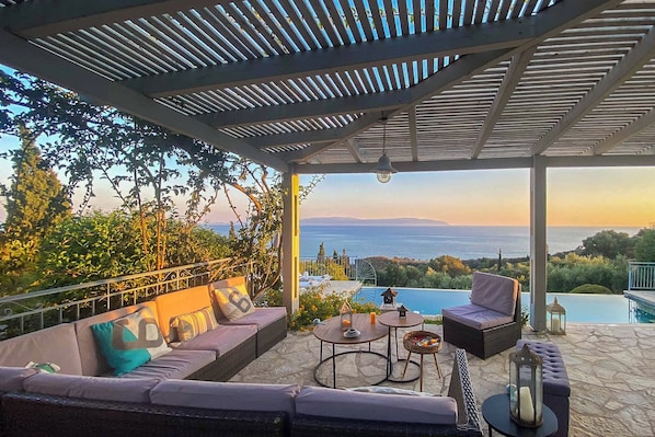 A covered outdoor seating area with a view of the sea, a pool, and lush greenery.
