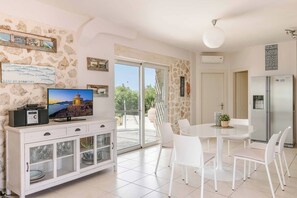 A modern living room with a television, white furniture, a dining table, and a view through a glass door.
