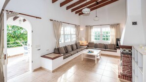 Un salon spacieux avec un canapé, une table basse et une cheminée. De grandes fenêtres, ornées de rideaux, offrent une vue sur la verdure environnante.