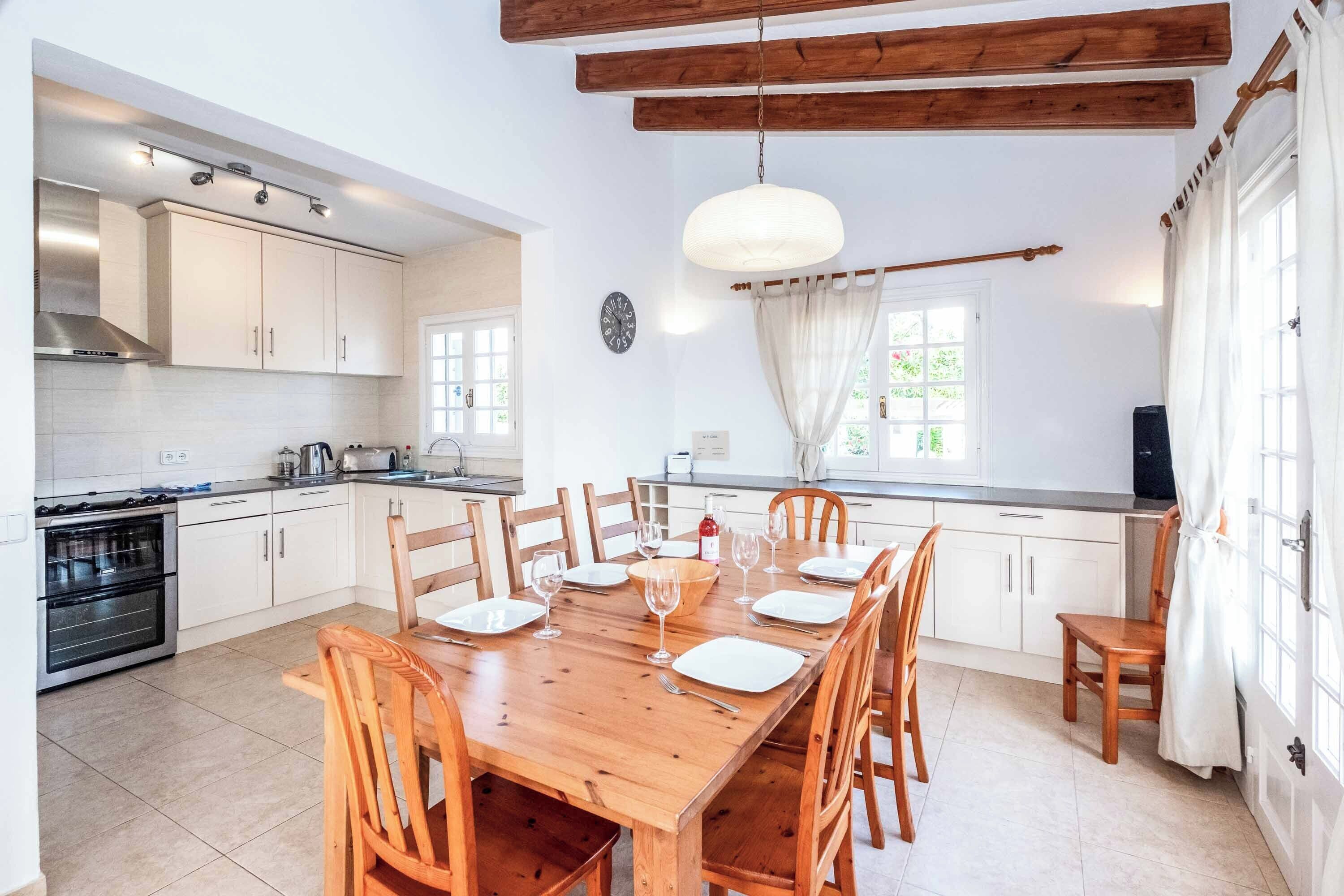 A modern kitchen with white cabinets, a wooden dining table set for six, and a large window with curtains.