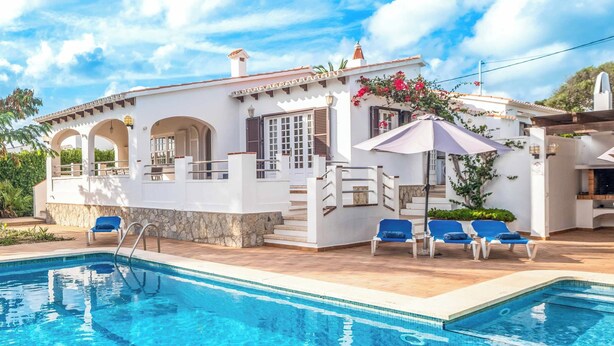 Une villa blanche avec une piscine, des fauteuils de jardin bleus et une pergola surmontée d’un parasol violet.