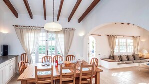 Un espace repas comprenant une table et des chaises en bois, un luminaire suspendu et une vue sur l’extérieur grâce à de grandes fenêtres.