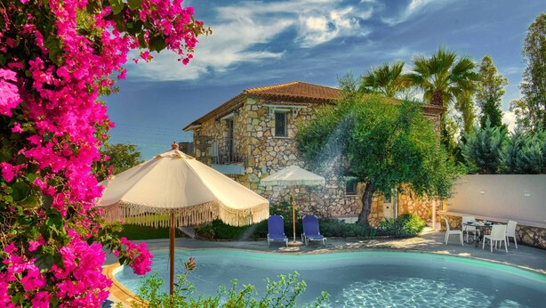 A stone villa with a swimming pool, surrounded by lush greenery and pink flowers.