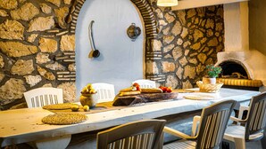 A stone-walled room with a rustic fireplace, a wooden table set with bread and tomatoes, and a hanging light fixture.