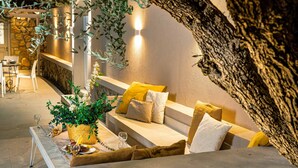A cozy seating area with a stone wall, a large tree trunk, and a table set with a bottle, glasses, and a basket of fruit.