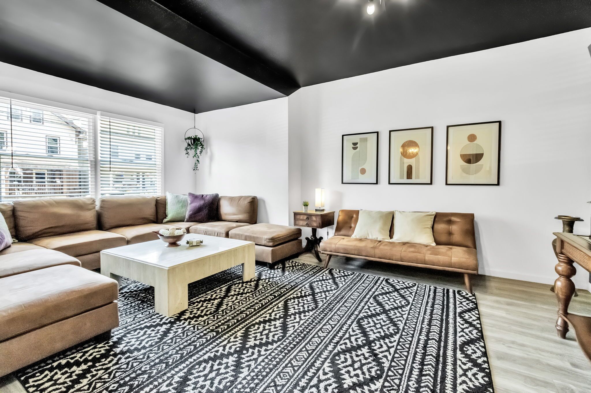 A modern living room with a sectional sofa, a coffee table, and a patterned rug. There are framed artworks on the wall and a plant hanging from the ceiling.