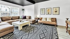Una sala de estar moderna con un sofá modular, una mesa de centro y un tapete estampado. Hay obras de arte enmarcadas en la pared y una planta colgando del techo.