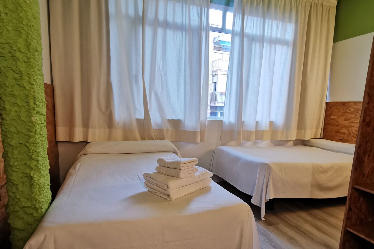 A room with two beds, a window with curtains, and a stack of towels on the bed.