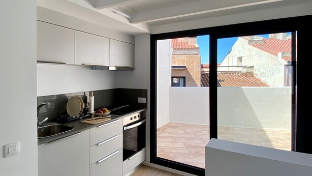 A modern kitchen with white cabinets, a black countertop, and a large sliding glass door leading to an outdoor patio. - Precioso Ático con Terraza Reformado (Mataró)