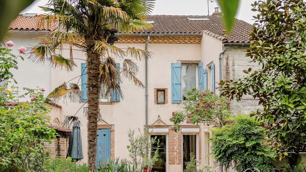 Une maison à deux étages avec un jardin, un palmier et un banc en métal blanc.