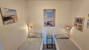 Una habitación pequeña y acogedora con dos camas individuales, un ventanal con vista a la playa, y decoración de paredes que representa escenas playeras y vida marina.