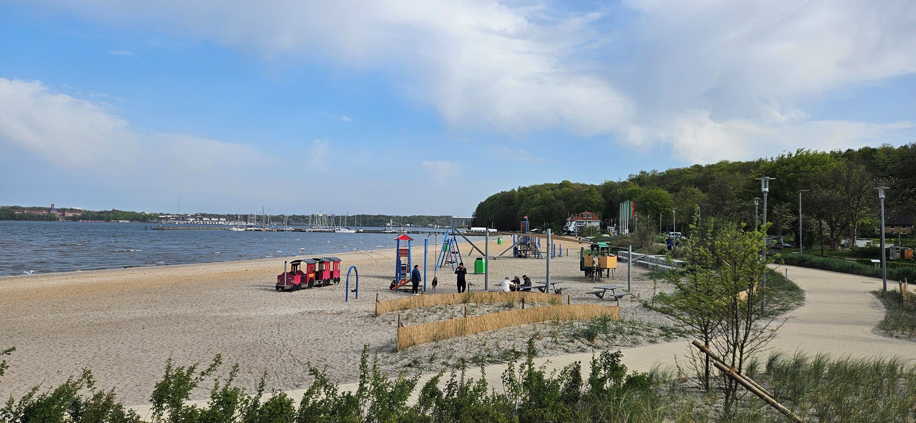 Uma praia com um pequeno trem vermelho, equipamentos de parque infantil e um caminho que leva a uma área de cais.