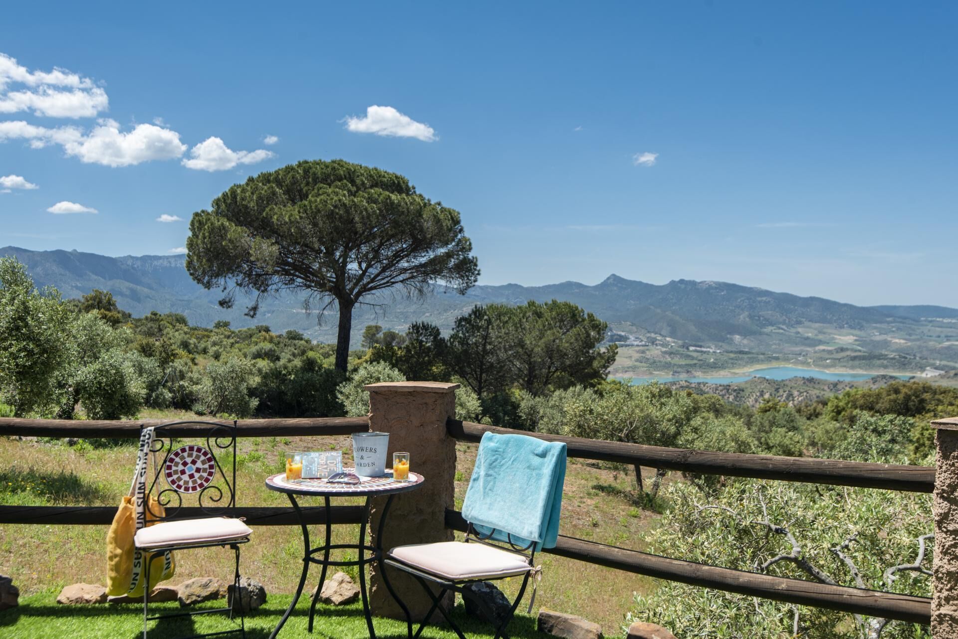 Eine Terrasse mit Tisch und zwei Stühlen, ein blaues Handtuch auf einem Stuhl sowie Blick auf Berge und einen See.