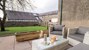 An outdoor seating area with wicker furniture, a table with a plate of croissants and glasses of orange juice, and a stone wall in the background. - Natland Hall Cottage (Kendal)