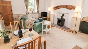 A cozy living room with a green sofa, a dining table set for two, a fireplace, and a television. - Natland Hall Cottage (Kendal)