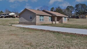 A single-story house with a brick exterior and a sloped roof, surrounded by a grassy area and other houses in the background. - Harris County Ranch Hideaway (Hamilton)