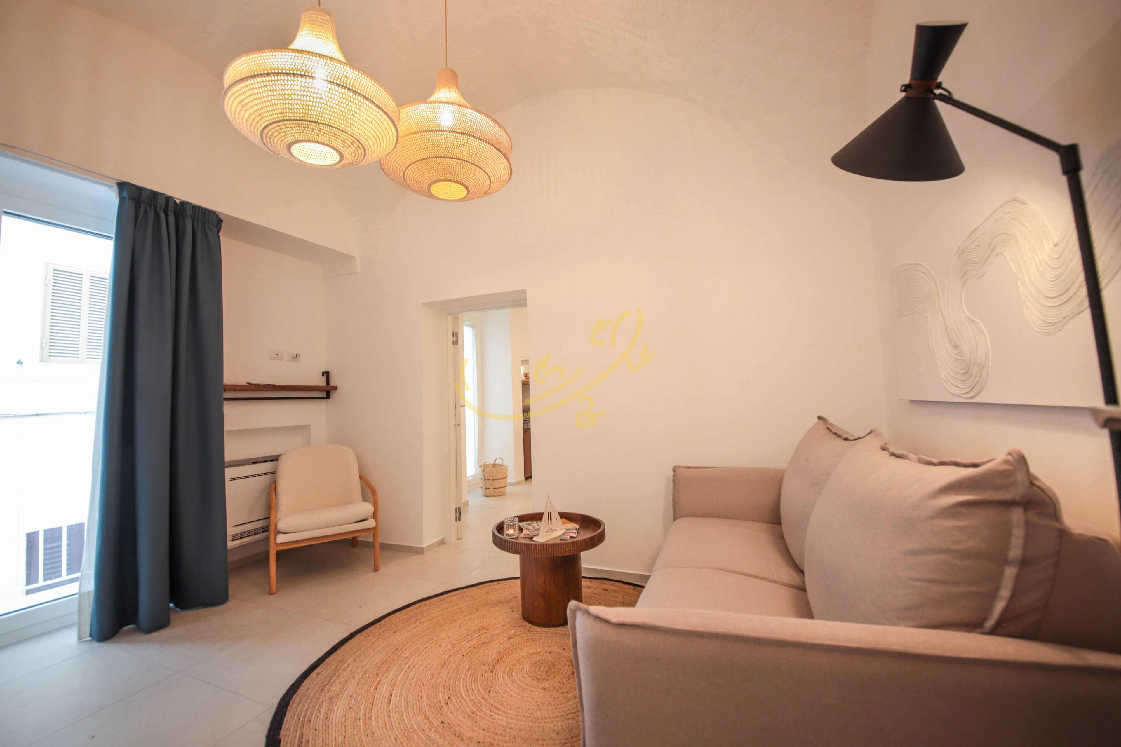 Una sala moderna con un sofá, una mesa de centro y una silla. Se ve una ventana con cortinas, una lámpara y una pared decorada.