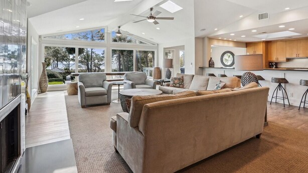 A spacious living area with a large sofa, two armchairs, and a coffee table. There is a kitchen area with wooden cabinets and a dining table. The room has large windows offering a view of trees and a building. - Oceanfront Sea Pines Home w/ Pool (Hilton Head Island)