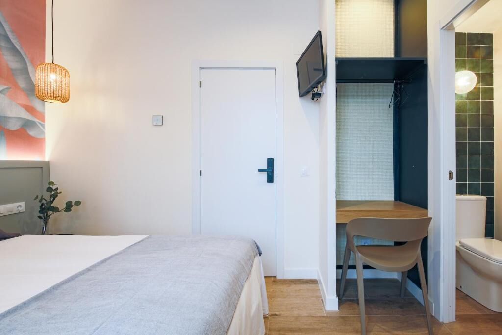 Un dormitorio con cama, escritorio y silla. Hay un televisor montado en la pared, una puerta y una ventana con persianas.