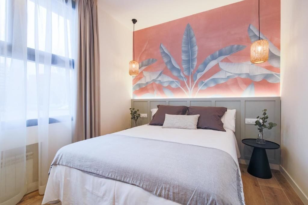 Una cama bien tendida con edredón blanco y gris, un gran mural de hojas en la pared y dos lámparas de noche.